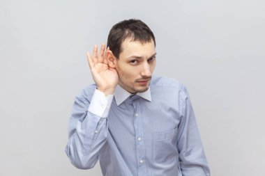 Meraklı yakışıklı bir adamın portresi, elinde bir şeyle durup bir şeyler dinliyor, özel sohbet ediyor, kameraya bakıyor, açık mavi gömlek giyiyor. Gri arka planda izole edilmiş kapalı stüdyo görüntüleri.