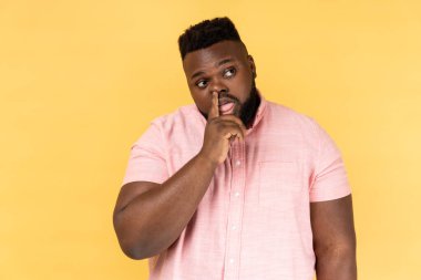 Misconduct, bad manners. Portrait of man wearing pink shirt picking nose and sticking out tongue with dumb comical expression, fooling around. Indoor studio shot isolated on yellow background.