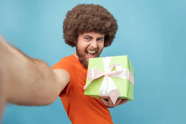 Portrait of happy man with Afro hairstyle wearing orange T-shirt standing with present box, showing gift, taking selfie, POV, being in festive mood. Indoor studio shot isolated on blue background.