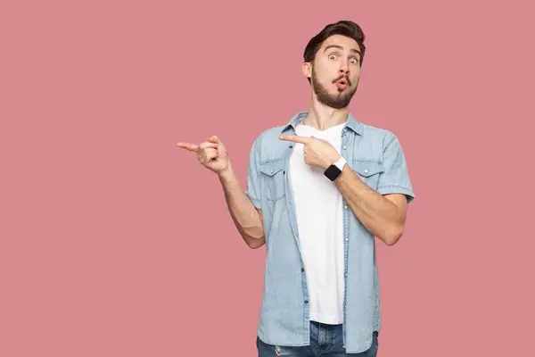 Amazed man in blue casual style shirt standing points away on copy space, discusses amazing promo, give way or direction, has astonished look. Indoor studio shot isolated on pink background