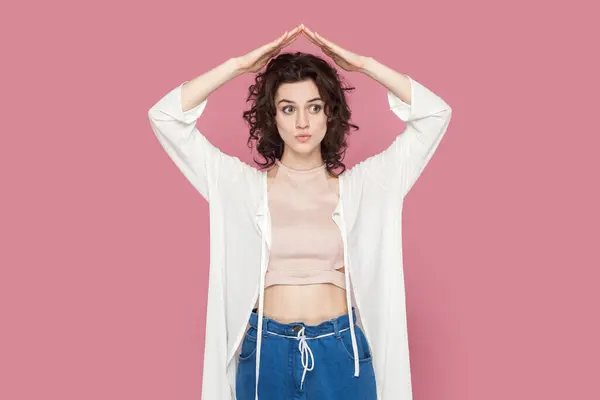 Portrait of beautiful young adult woman with curly hair wearing casual style outfit making roof with hands above her head, feels in safety. Indoor studio shot isolated on pink background.