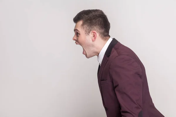 Side view portrait of aggressive annoyed handsome young man arguing with somebody, screaming with hate and anger, wearing violet suit and white shirt. Indoor studio shot isolated on grey background.