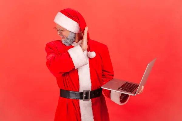 I don\'t want to look at this. Elderly man with gray beard wearing santa claus costume turning his face from computer, shocked content. Indoor studio shot isolated on red background.
