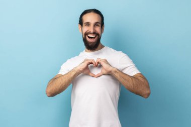 İmza atmana bayılıyorum. Benim sevgilim ol. Sakallı, beyaz tişörtlü romantik bir adamın portresi, kalp şeklinde, aşkla itiraf ediyor, kameraya bakıyor. Mavi arka planda kapalı stüdyo çekimi.