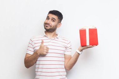Çizgili tişört giyen mutlu sakallı, mutlu bir adamın portresi kırmızı hediye kutusunu tutuyor. Gri arka planda izole edilmiş kapalı stüdyo görüntüleri.
