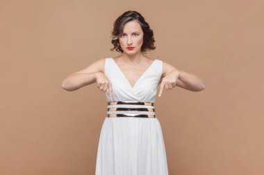 Burada ve şimdi. Kadın kendini beğenmiş bir şekilde işaret ediyor, derhal itaat talep ediyor, otoritesini gösteriyor, beyaz elbise giyiyor. Açık kahverengi arka planda kapalı stüdyo çekimleri.