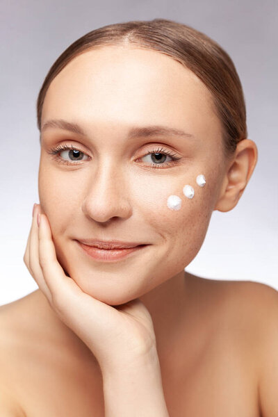 Крупный план портрета улыбающейся красивой женщины с голыми плечами, накладывающей крем на щеку, смотрящей в камеру с рукой под подбородком, счастливым выражением лица. Крытая студия снята на сером фоне.