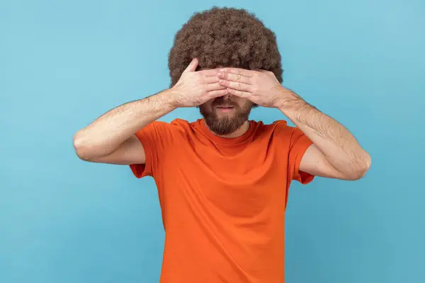 手でオレンジ色のTシャツの目を閉じたアフロヘアスタイルの男性の肖像画は ストレスの多い状況から隠れて 問題を無視して それを見たくありません ブルーの背景に隔離された屋内スタジオショット — ストック写真