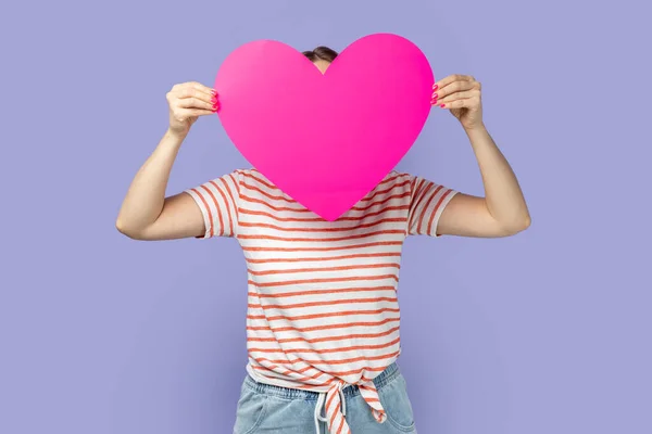 Portrait of shy unknown blond woman wearing striped T-shirt holding big pink heart and hiding her face behind love symbol, anonymous feelings. Indoor studio shot isolated on purple background.