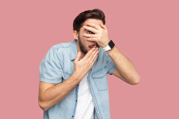 Portrait of scared frightened bearded man in blue casual style shirt standing peeks through fingers, hides face, stares with at something spooky. Indoor studio shot isolated on pink background.
