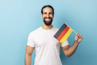 Almanya bayrağını tutan beyaz tişörtlü, sakallı, olumlu gülümseyen bir adamın portresi. 3 Ekim 'de Almanya bayrağını taşıyan, kameraya bakan. Mavi arka planda kapalı stüdyo çekimi.