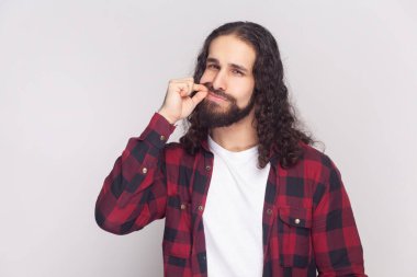 Kareli kırmızı gömlekli uzun kıvırcık saçlı yakışıklı sakallı adamın portresi sır olarak kalıyor, ağzını fermuarını çekip kameraya bakıyor. Gri arka planda izole edilmiş kapalı stüdyo görüntüleri.