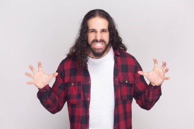 Kızgın sakallı, uzun kıvırcık saçlı, uzun, kırmızı gömlekli bir adamın portresi öfkeyle hareket ediyor, birine bağırıyor, suratını asıyor. Gri arka planda izole edilmiş kapalı stüdyo görüntüleri.