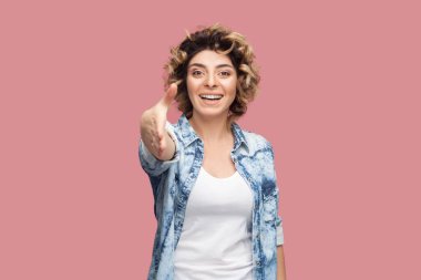 Neşeli, iyimser, kıvırcık saçlı, mavi gömlekli, elini uzatıp selam veren güzel bir kadının portresi. Pembe arka planda kapalı stüdyo çekimleri.
