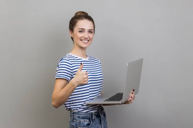 Çizgili tişört giyen, elinde dizüstü bilgisayarla ayakta duran, tele-çalışmaktan memnun bir kadın, işini seviyor. Gri arka planda izole edilmiş kapalı stüdyo görüntüleri.