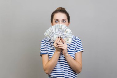 Yüzünün yarısını bir sürü banknotla kaplayan çizgili tişört giyen, büyük gözlü kameraya bakan şaşkın kadının portresi. Gri arka planda izole edilmiş kapalı stüdyo görüntüleri.