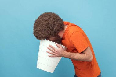 Afro saçlı, turuncu tişörtlü, kötü hislerden muzdarip, mide rahatsızlığı olan hasta, bozuk ürün olan bir adamın yan görüntüsü. Mavi arka planda kapalı stüdyo çekimi.
