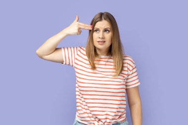 Çizgili tişört giyen sarışın bir kadının portresi. Parmaklarını kafasının yanında tutuyor. Taklit tabanca, panik stresli ifadeyle kameraya bakıyor. Mor arka planda izole edilmiş kapalı stüdyo çekimi.