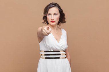 Katı, orta yaşlı, dalgalı saçlı, kameraya işaret eden, ciddi şekilde patronluk taslayan, beyaz elbise giyen bir kadın. Açık kahverengi arka planda kapalı stüdyo çekimleri.