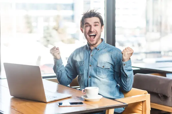 穿着蓝色牛仔裤衬衫的快乐而积极的自由职业者在笔记本电脑上工作的画像 紧握拳头 庆祝他的成功 看着相机 在大窗户附近的室内拍摄 咖啡馆背景 — 图库照片