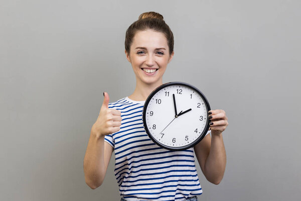 Portrait of smiling delighted positive woman with bun hairstyle wearing striped T-shirt standing with wall clock in hands, showing thumb up. Indoor studio shot isolated on gray background.