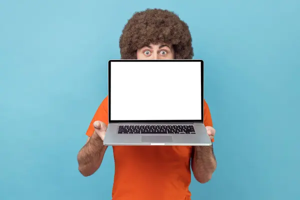 Anonymous man with Afro hairstyle in orange T-shirt holding laptop in hands, hiding half of his face, copy space for promotion, looking at camera. Indoor studio shot isolated on blue background.