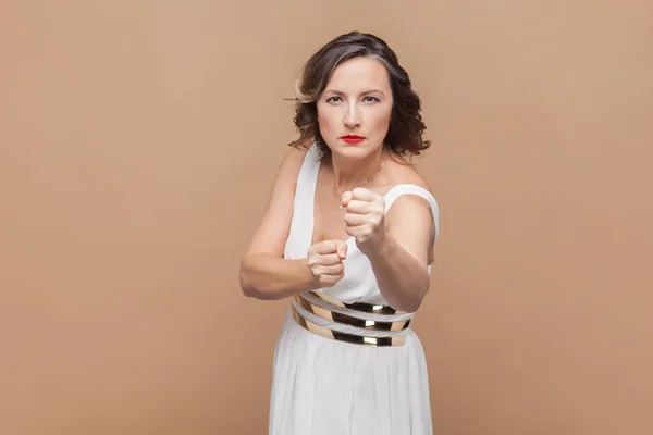 怒っている中年女性の肖像画 ひび割れた拳で 誰かと戦い 怒りでボクシングし 白いドレスを着ています 明るい茶色の背景で隔離された屋内スタジオショット — ストック写真