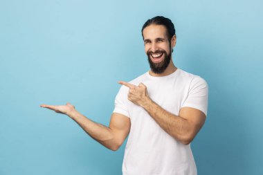 Beyaz tişörtlü, sakallı, tatmin olmuş bir adamın portresi. Ticari metinler için avuç içinde fotokopi alanı sunuyor. Fikir sunumlu boş bir duvar. Mavi arka planda kapalı stüdyo çekimi.