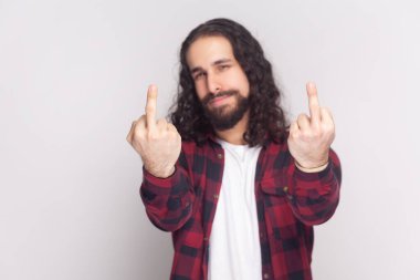 Fuck off. Portrait of displeased bearded man with long curly hair in checkered red shirt shows fuck sign, showing his negative feeling. Indoor studio shot isolated on gray background.