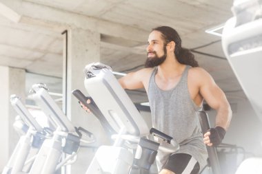 Uzun kıvırcık saçlı, spor giyimli spor kıyafetleri giymiş koşu bandında koşan vücut, vücut ve sağlık hizmetlerinden önce kardiyo ısınması yaşayan kaslı bir adamın yan görüntüsü. Kapalı çekim.