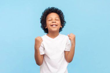 Son derece mutlu, iyimser, kıvırcık saçlı, gözleri kapalı, yumruklarını sıkarak okuldaki başarısını kutlayan bir çocuğun portresi. Mavi arka planda kapalı stüdyo çekimi.