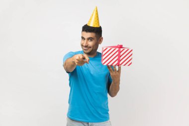 Senin için bir hediye. Dost canlısı, tıraşsız, mavi tişörtlü ve sarı parti konisi giymiş, elinde hediye kutusuyla kameraya bakan yakışıklı bir adam. Gri arka planda izole edilmiş kapalı stüdyo görüntüleri.