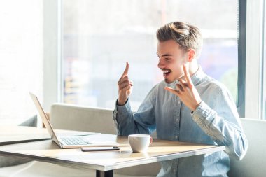 Heyecanlı, sakallı, serbest çalışan genç bir adamın portresi kafede oturuyor ve internette rock 'n roll jestleri hakkında konuşuyor, kot ceket giyiyor. Büyük pencereye karşı kapalı çekim.