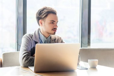 Düşünceli, düşünceli, sakallı, serbest çalışan genç bir adamın portresi kafede oturuyor ve dizüstü bilgisayarda çalışıyor, yeni projesini düşünüyor, gri ceket giyiyor. Büyük pencereye karşı kapalı çekim.