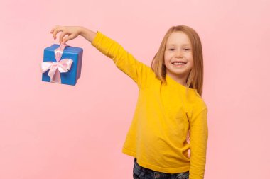 Mavi ambalajlı, mutlu bir ifadeyle kameraya bakıp sarı kazak giyen neşeli sarışın küçük kızın portresi. Pembe arka planda kapalı stüdyo çekimleri.