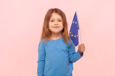 Avrupa Birliği bayrağını tutan neşeli küçük bir kızın portresi, kameraya bakıyor, Avrupa ülkesine gidiyor, mavi süveter giyiyor. Pembe arka planda kapalı stüdyo çekimleri.