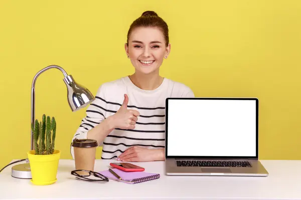 Smiling happy young woman employee pointing empty laptop screen, mock up display for advertisement, showing thumb up, website promotion. Indoor studio studio shot isolated on yellow background.