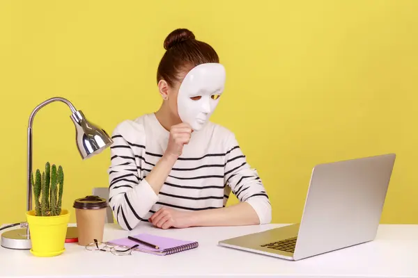 Unknown anonymous woman covering her face with white mask, hiding personality while sitting on workplace with notebook. Indoor studio studio shot isolated on yellow background.