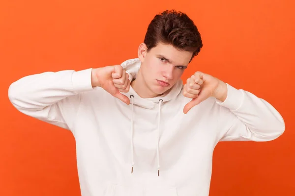 Portrait of young man wearing white hoodie expresses dislike with body language, keeps thumb down and frowns face, has displeased face. Indoor studio shot isolated on orange background.