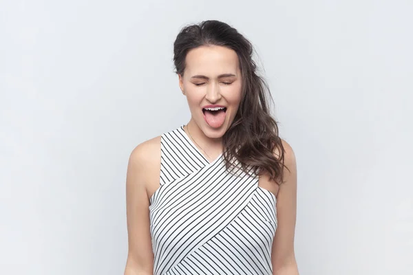 stock image Portrait of funny positive brunette woman standing with closed eyes, showing tongue out, demonstrates childish behavior, wearing striped dress. Indoor studio shot isolated on gray background.