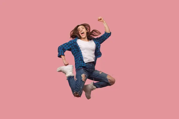 全长肖像极度快乐的棕色头发女人跳跃与紧握拳头庆祝她的成功的成就 穿着格子衬衫 在粉色背景下被隔离的室内拍摄 — 图库照片