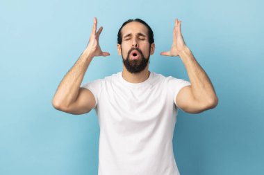Portrait of man with beard wearing white T-shirt touching his head and showing explosion, looking worried and shocked, deadline, professional burnout. Indoor studio shot isolated on blue background. clipart
