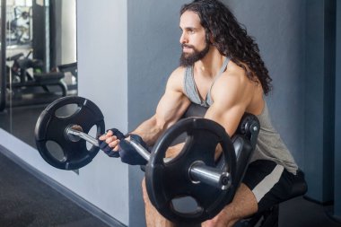 Uzun kıvırcık saçlı, spor kıyafetli güçlü bir adamın portresi spor salonunda vücudunu çalıştırıyor, halter kaldırıyor, kollar ve omuzlar için egzersiz yapıyor. Kapalı çekim.