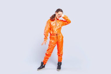 Full length portrait of hip-hop dancer teenage girl with brunette hair wearing bright orange jumpsuit standing showing victory sign. Indoor studio shot isolated on gray background. clipart