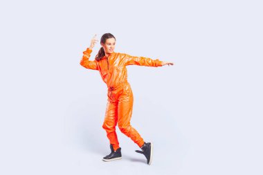 Uzun boylu, gülümseyen hip-hop dansçısı esmer saçlı, parlak turuncu tulumlu kollarını kaldırmış dans eden genç kız portresi. Gri arka planda izole edilmiş kapalı stüdyo görüntüleri.