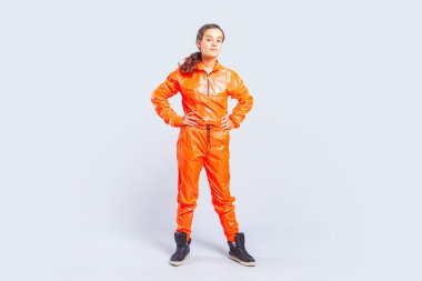 Full length portrait of confident hip-hop dancer teenage girl with brunette hair wearing bright orange jumpsuit standing with hands on waist. Indoor studio shot isolated on gray background. clipart
