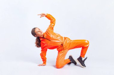 Parlak turuncu tulumlu, esmer, havalı hip-hop dansçısı genç bir kızın tam boy portresi performansını sunuyor. Gri arka planda izole edilmiş kapalı stüdyo görüntüleri.