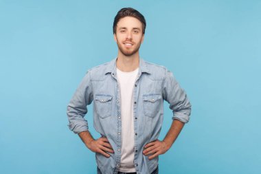 Gülümseyen, tüylü, kameraya bakan, elleri kalçasında duran, keyifli, kot gömlek giyen yakışıklı bir adamın portresi. Mavi arkaplanda kapalı çekim.