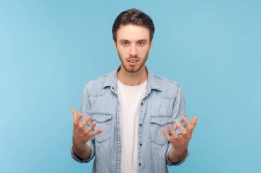 Öfke ve öfkeyle çığlık atan, kot gömlek giyen, kameraya bakan, saldırgan, tıraşsız bir adamın portresi. Mavi arkaplanda kapalı çekim.