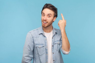 Tüylü neşeli adam ön parmağıyla işaret ediyor, parlak bir fikir elde etmek için işaret parmağını kaldırıyor, kameraya heyecanla bakıyor, kot gömlek giyiyor. Mavi arkaplanda kapalı çekim.